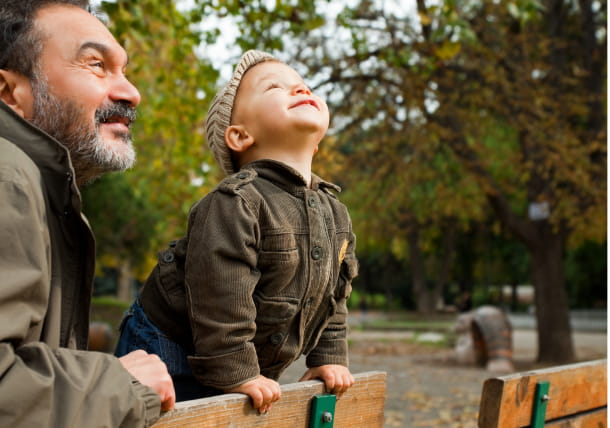 A father and his son are leaning on a bench in the park, looking up and smiling at the trees and sky.